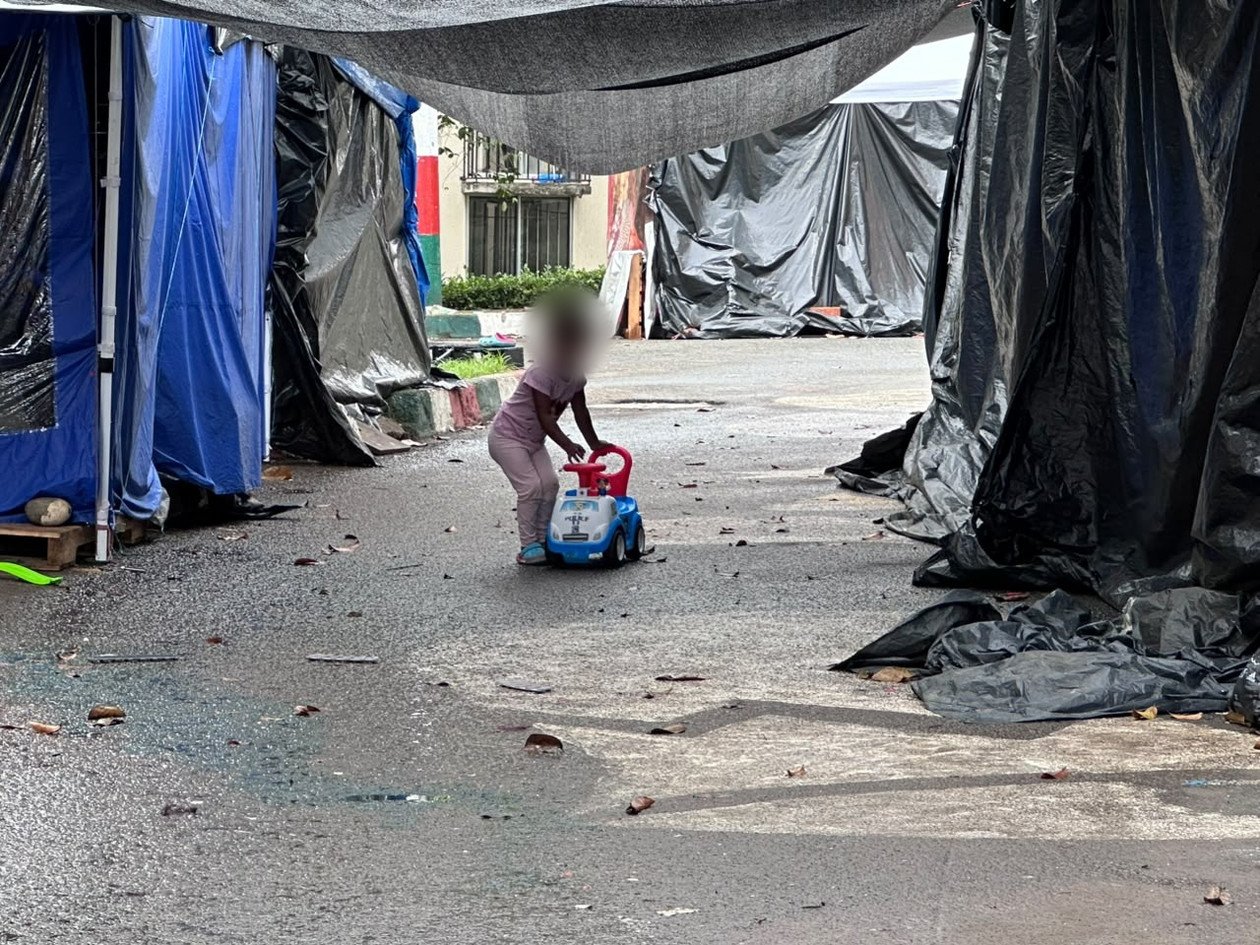 Una niña juega en un carro de juguete entre las carpas de Altos de Santa Elena, Cali (rostro desenfocado)