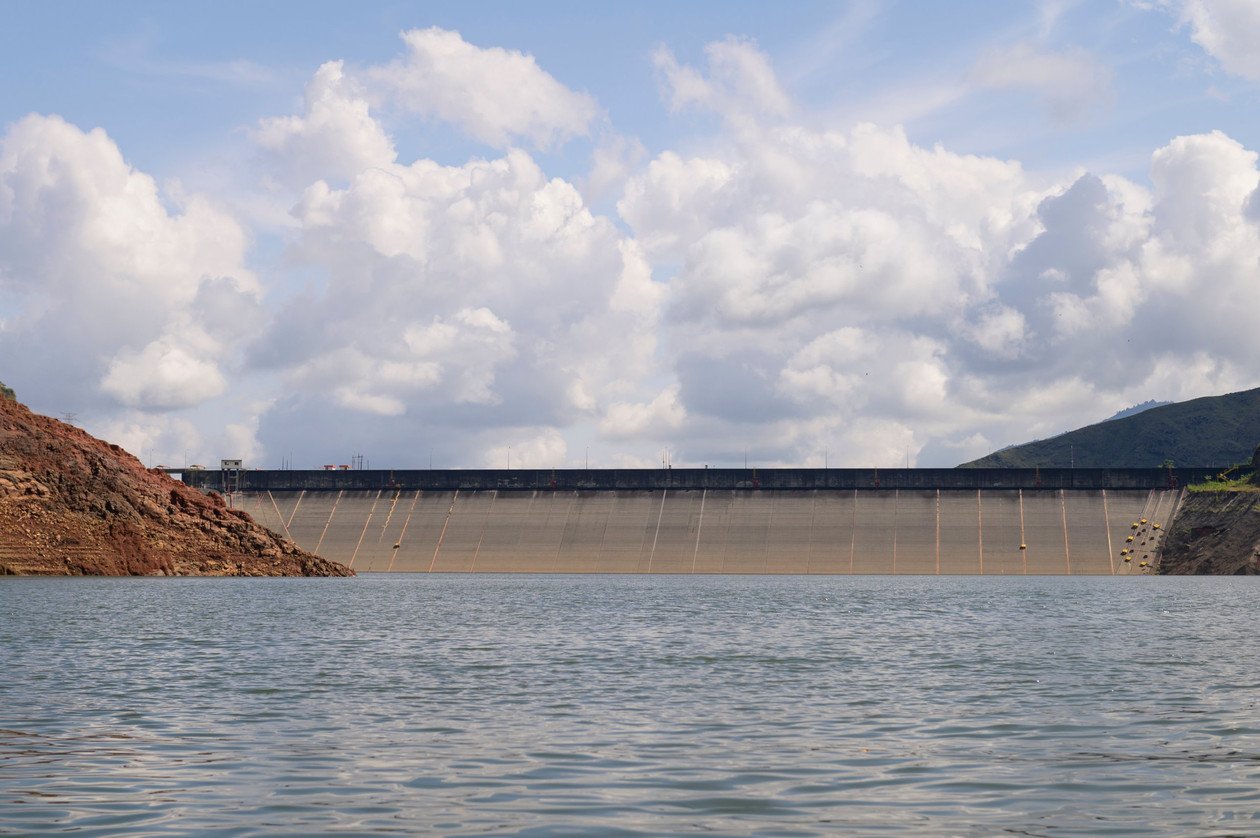 Vista de la presa de una hidroeléctrica desde el embalse, con montañas y nubes al fondo