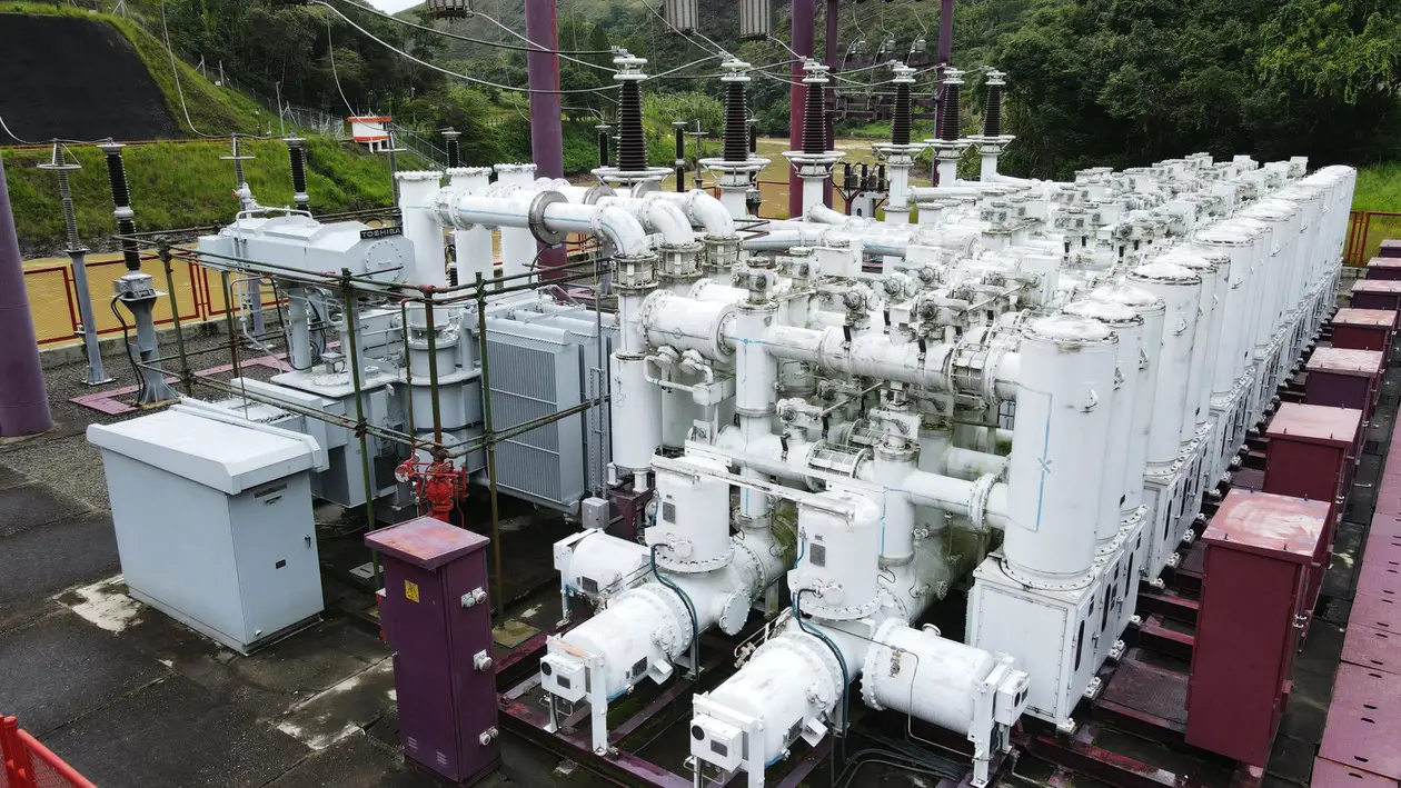 Equipos de una subestación eléctrica, con transformadores y tuberías blancas, junto a un río
