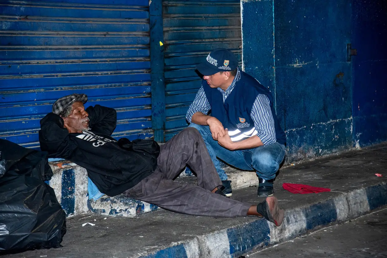 Funcionario de la Alcaldía de Cali conversa en cuclillas con un hombre recostado en un andén, de noche