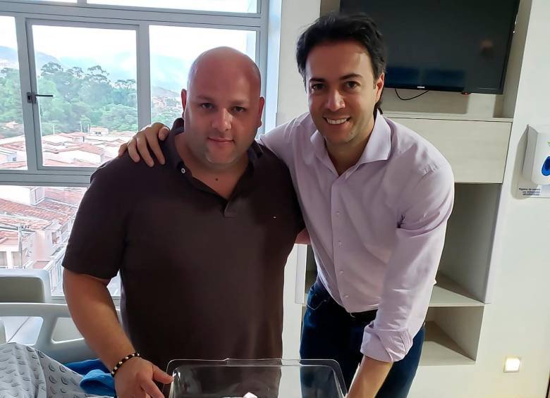 Dos hombres posan abrazados en una habitación con una ventana y un televisor