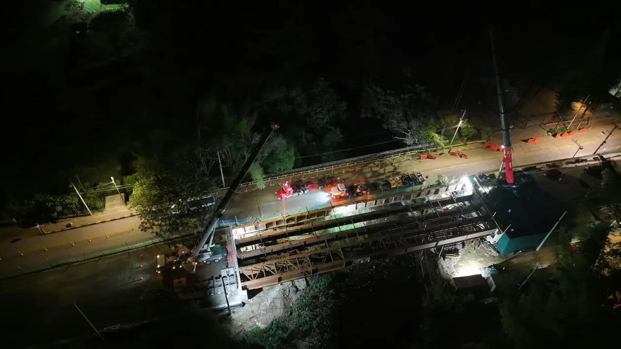 Vista aérea nocturna del puente sobre el río Pance, con las vigas metálicas, dos grúas y camiones sobre la vía